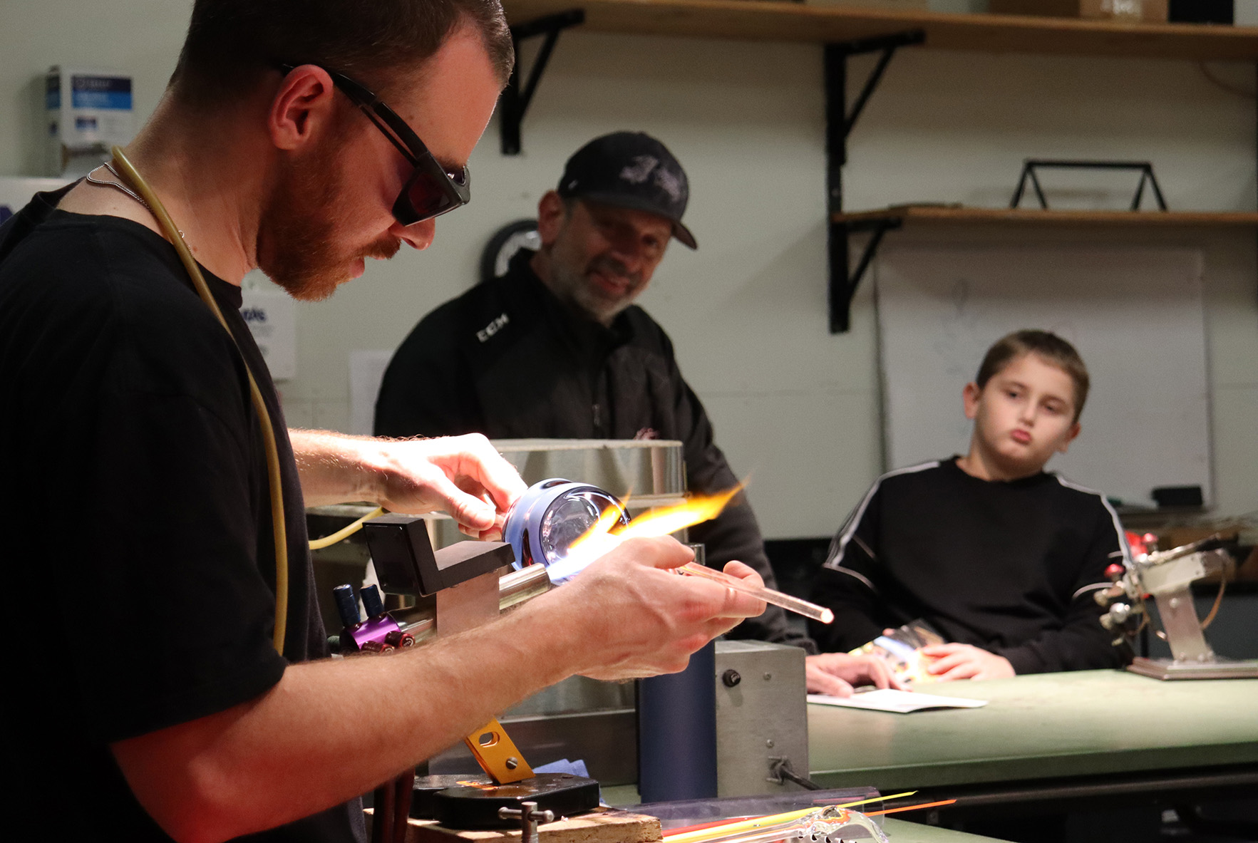 Glass artist working at a table while a father and son watch in the background