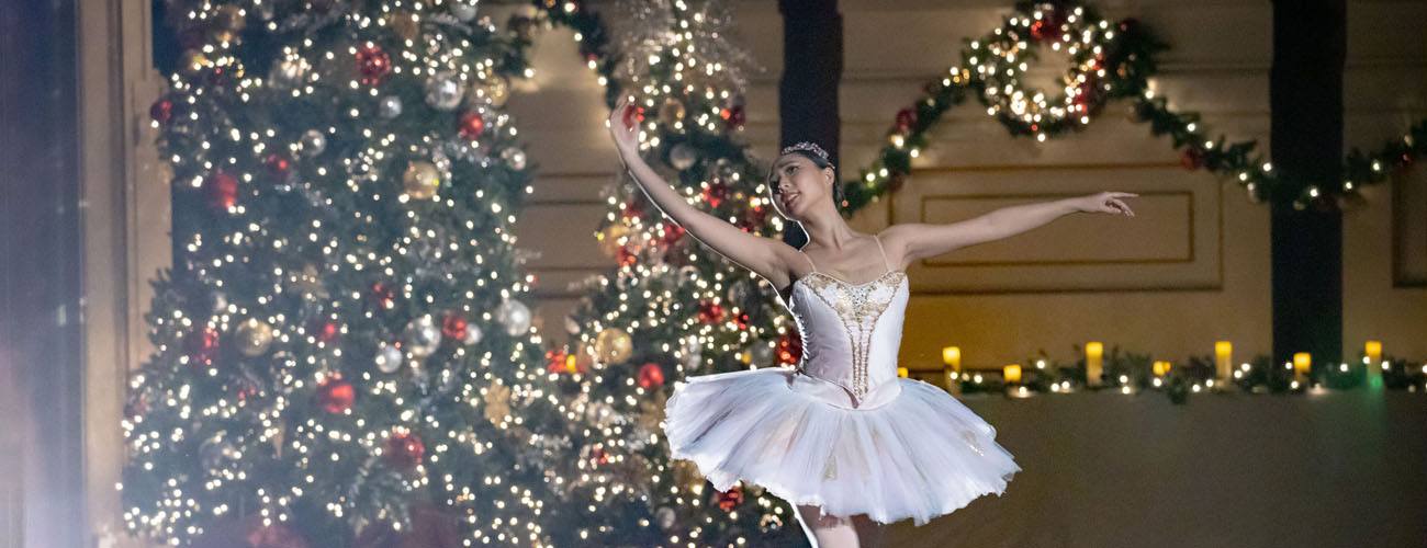 Ballerina in a white tutu dancing in front of a grand Christmas tree and holiday decorations.