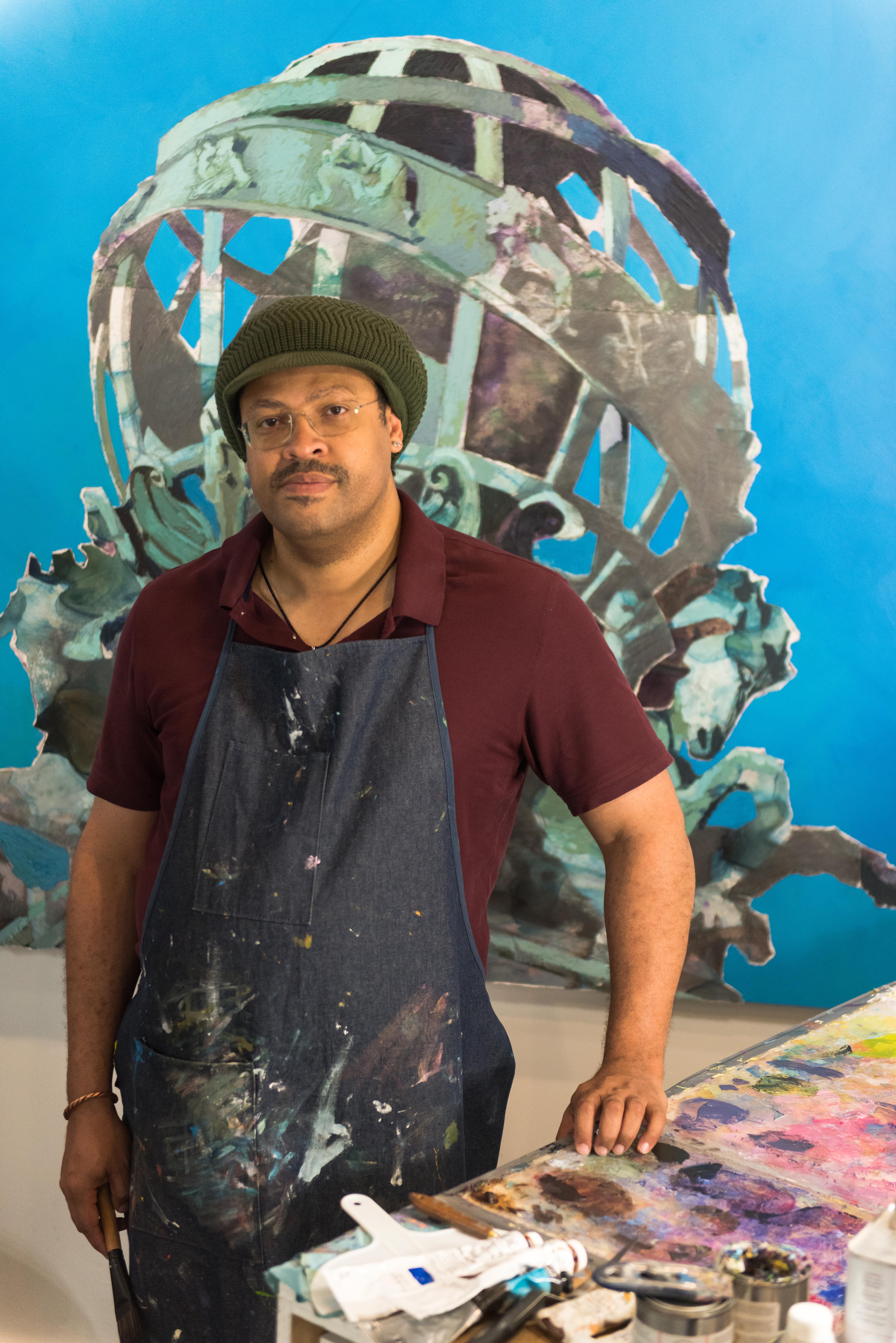 Artist Ramon Riley wearing an apron covered in paint, standing in front of one of his works of art.